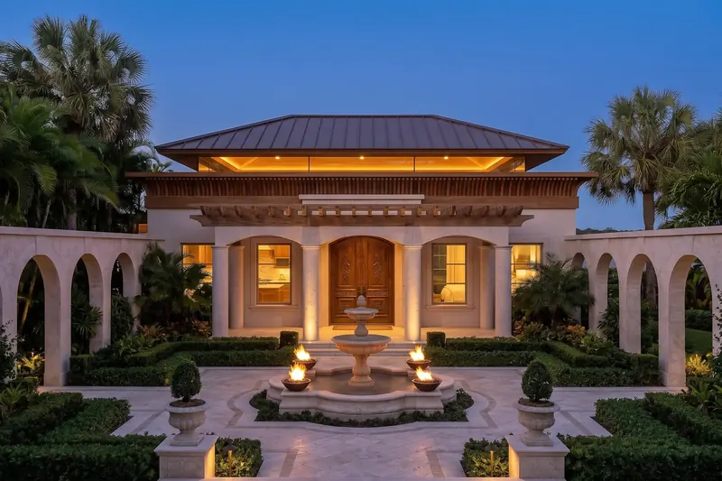 Classical pavilion home at dusk with arched colonnades, carved wood doors and a fountain ringed by fire bowls