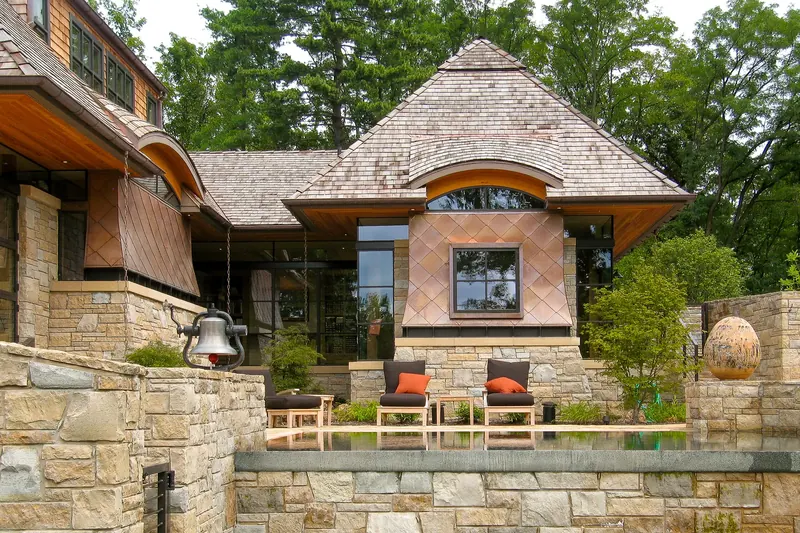 Stone and shingle home with diamond-pattern copper cladding, curved eyebrow dormer and a terrace with lounge chairs by a pool