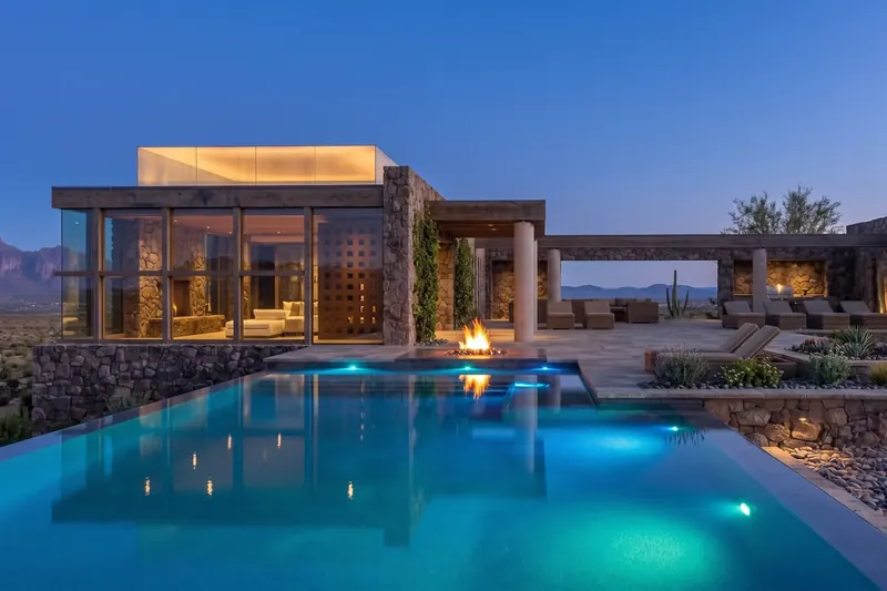 Stone and glass desert home at dusk with a glowing lantern roof, lit infinity pool, fire feature and mountain views