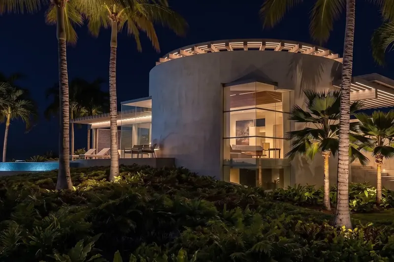 Curved white rotunda with a glass corner window and lit pergola at night, framed by palm trees and a glowing pool