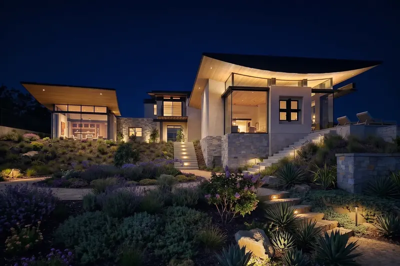 Hillside modern home at night with upswept wood-lined roofs, glowing glass rooms, stone base and lit stairs through gardens