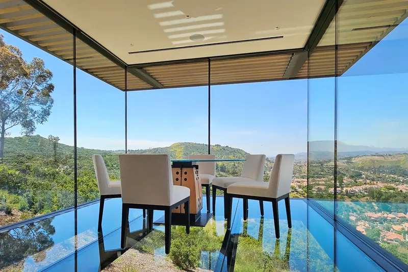 Glass-walled dining nook with a glass floor and round table, floating over green hills and a valley neighborhood