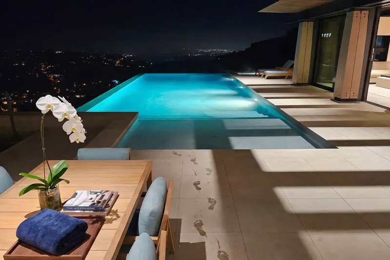 Night view of a lit infinity pool beside an outdoor dining table with an orchid, city lights in the valley below