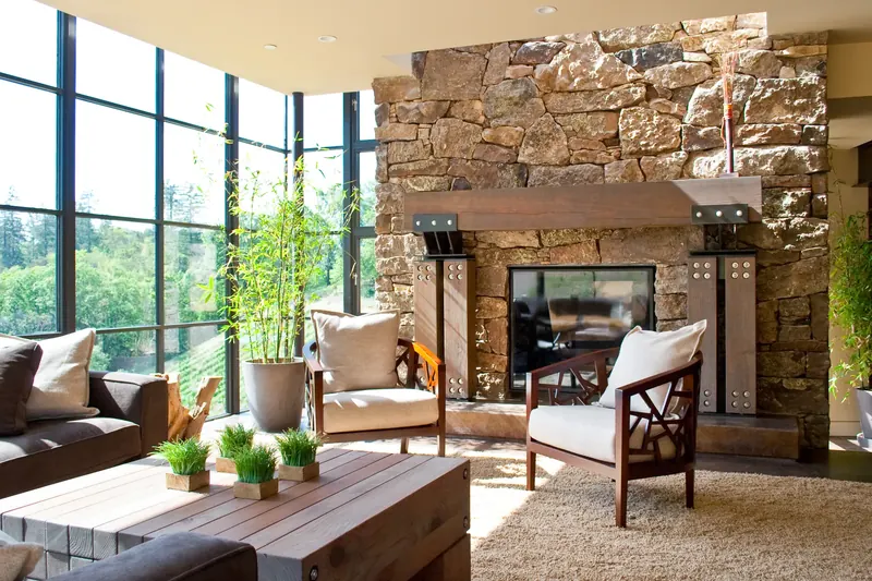 Sunlit living room with a rough stone fireplace, bolted timber-and-steel mantel, corner window wall and vineyard view