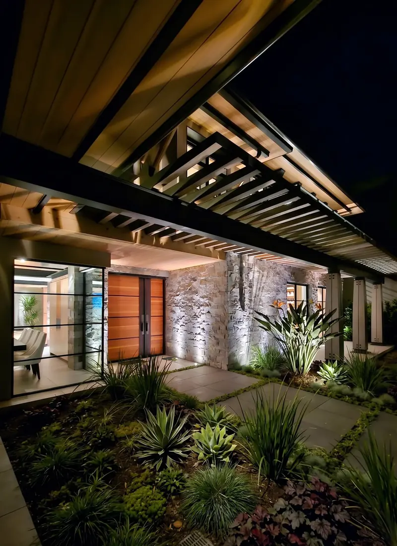 Entry at night with wood double door, uplit stone wall and slatted canopy over a lit garden path