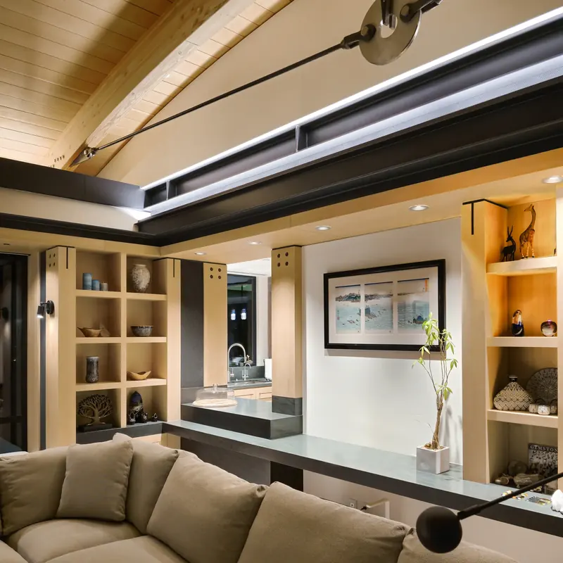 Built-in light wood shelving and stone counter beneath a lit steel beam and arched wood ceiling with tension rod