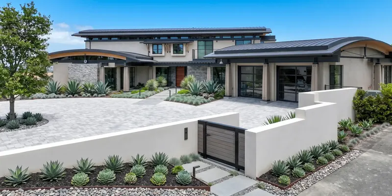 Front of the Arches House with curved dark metal roofs, stucco walls, stone accents and a gated agave and succulent garden