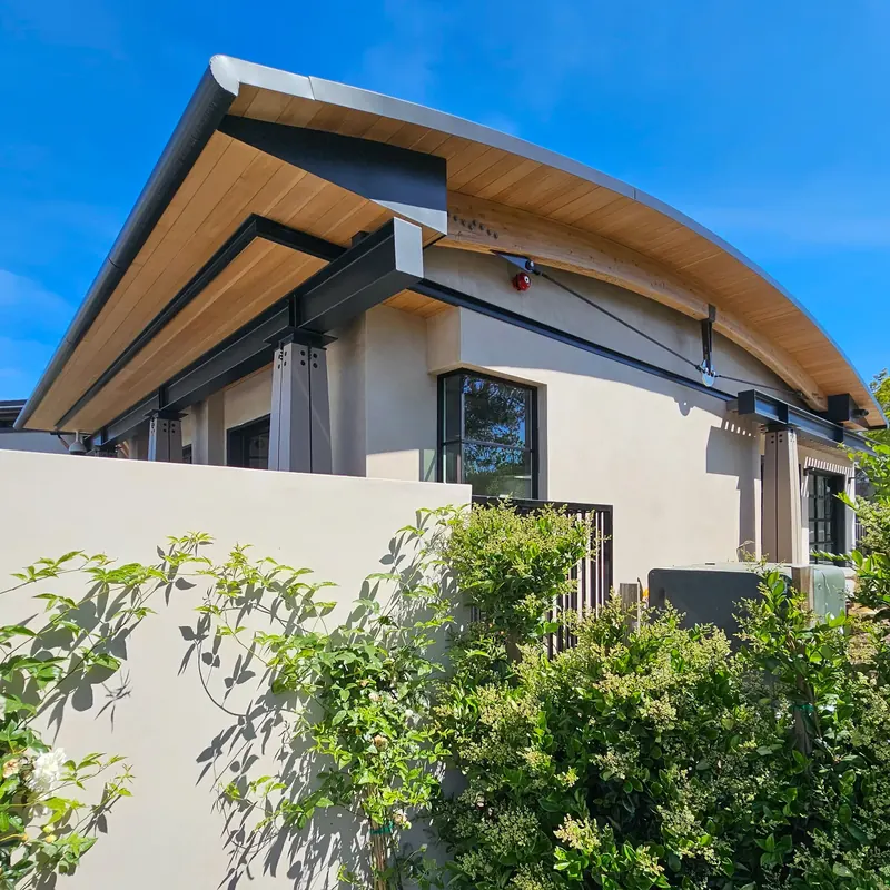 Sweeping curved roof with wood soffit, exposed wood arch and tension rod above stucco walls and leafy shrubs