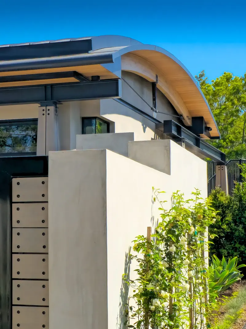 Stucco screen wall beside a bolted panel gate, beneath a curved roof edge with steel beams and a tension rod