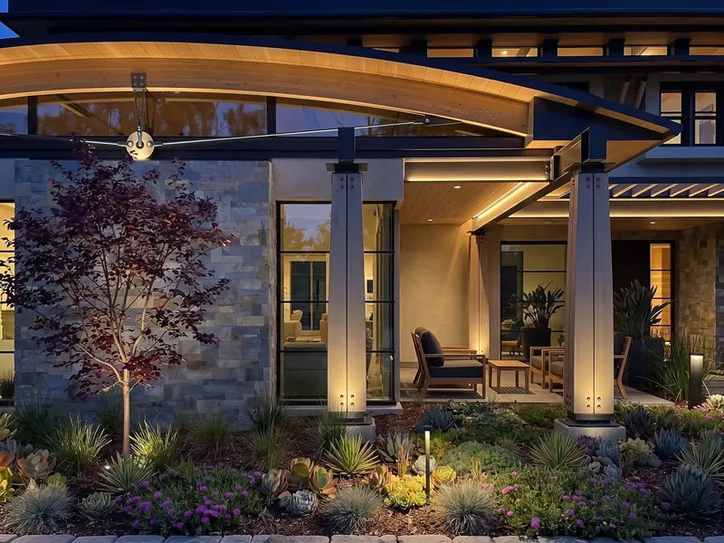 Arches House at dusk: a lit wood arch and steel tension rod above a stone wall, tall windows and a covered patio