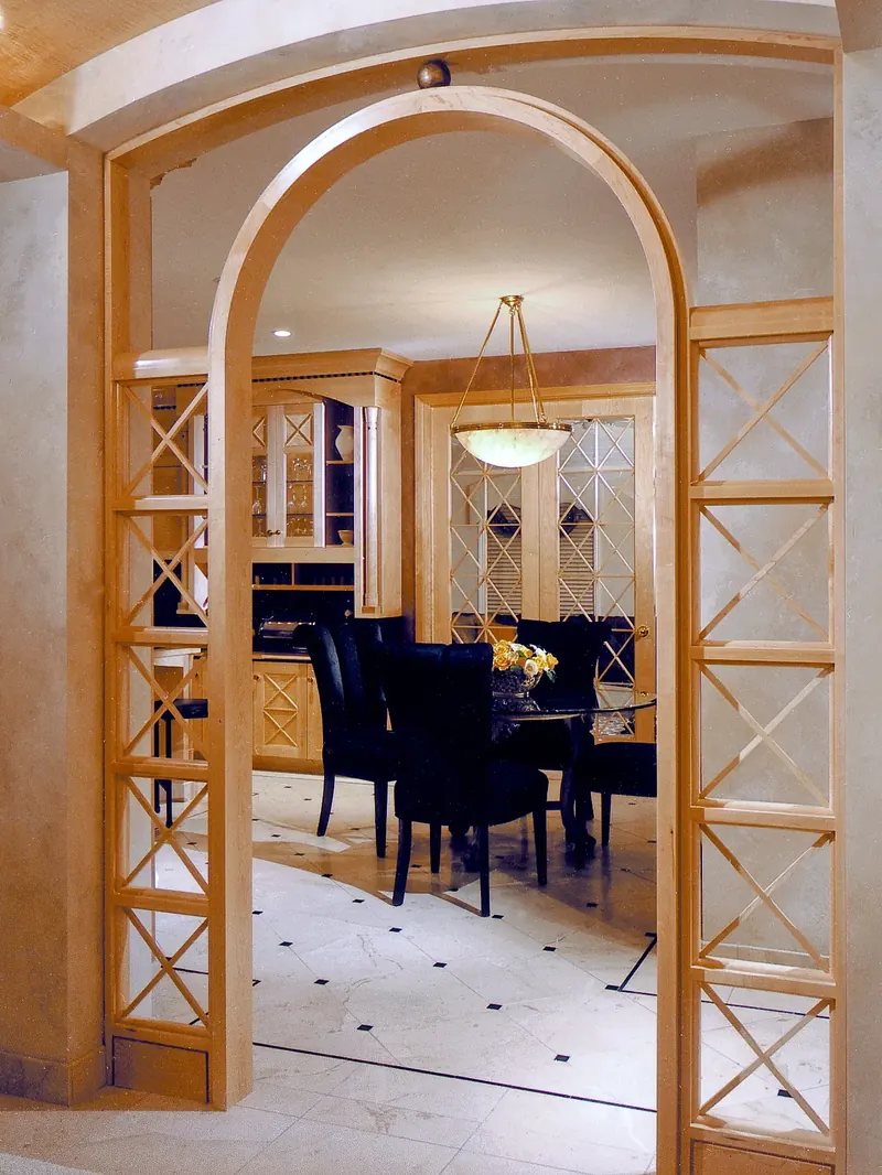 Wood archway with lattice side panels framing a round glass table, black chairs and kitchen cabinetry beyond
