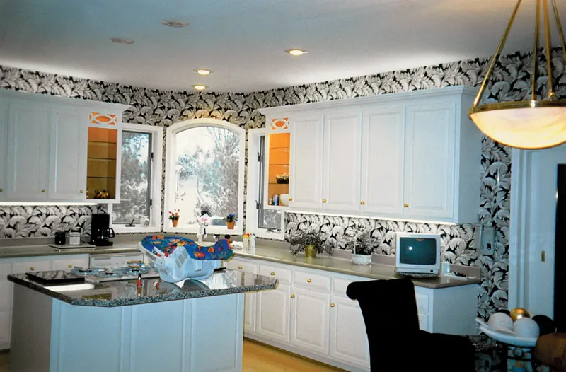 Kitchen before remodeling: white cabinets, bold leaf-pattern wallpaper, arched window and granite island
