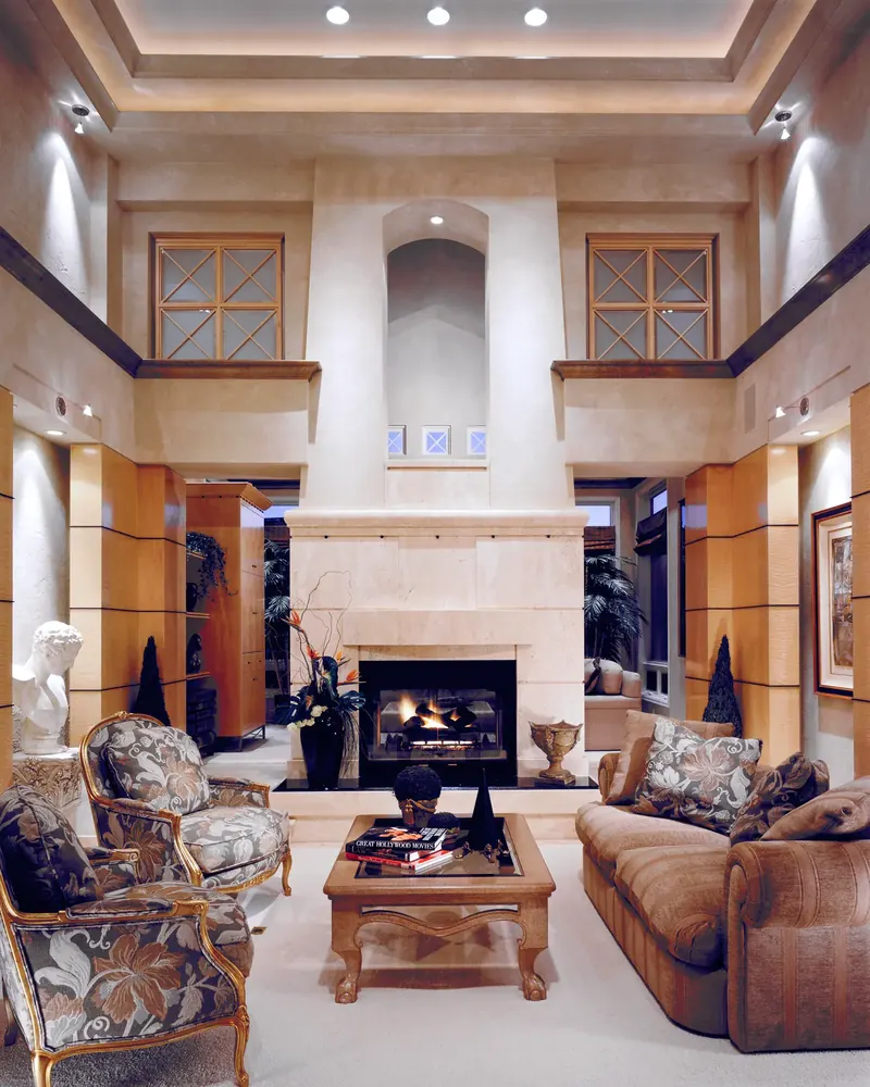 Two-story living room after remodeling, with a tall stone-clad fireplace, upper cross-pane windows and gilded armchairs