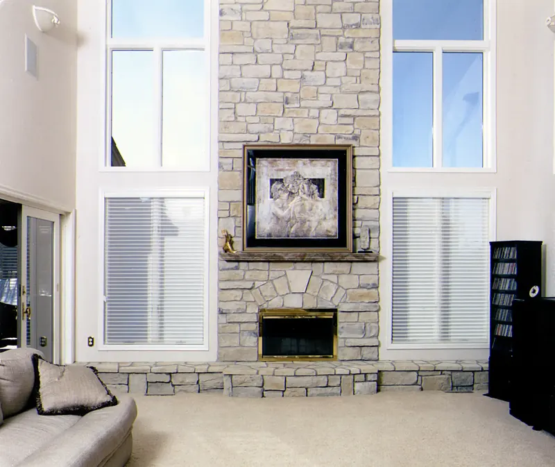 Living room before remodeling: stacked-stone fireplace between tall windows with blinds and plain white walls