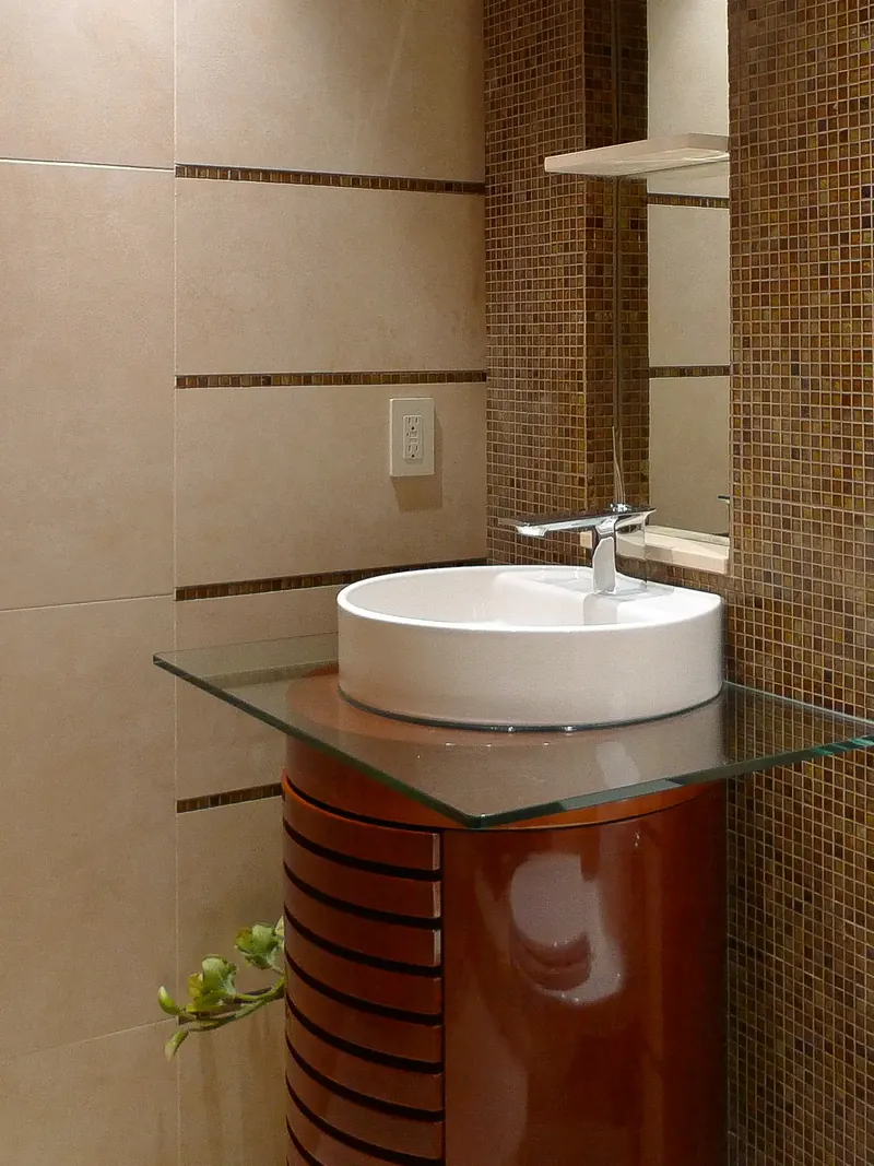 Cylindrical slatted wood vanity cabinet under a glass counter and round white vessel sink in a tiled bathroom