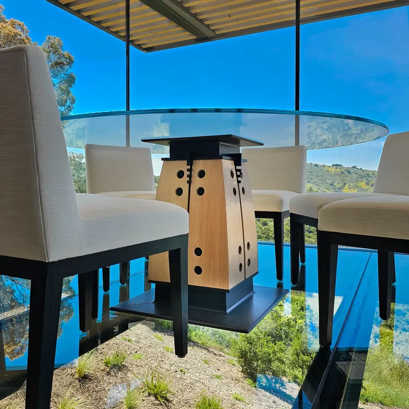 Round glass dining table on a tapered timber and black pedestal, with upholstered chairs and green hillside views beyond