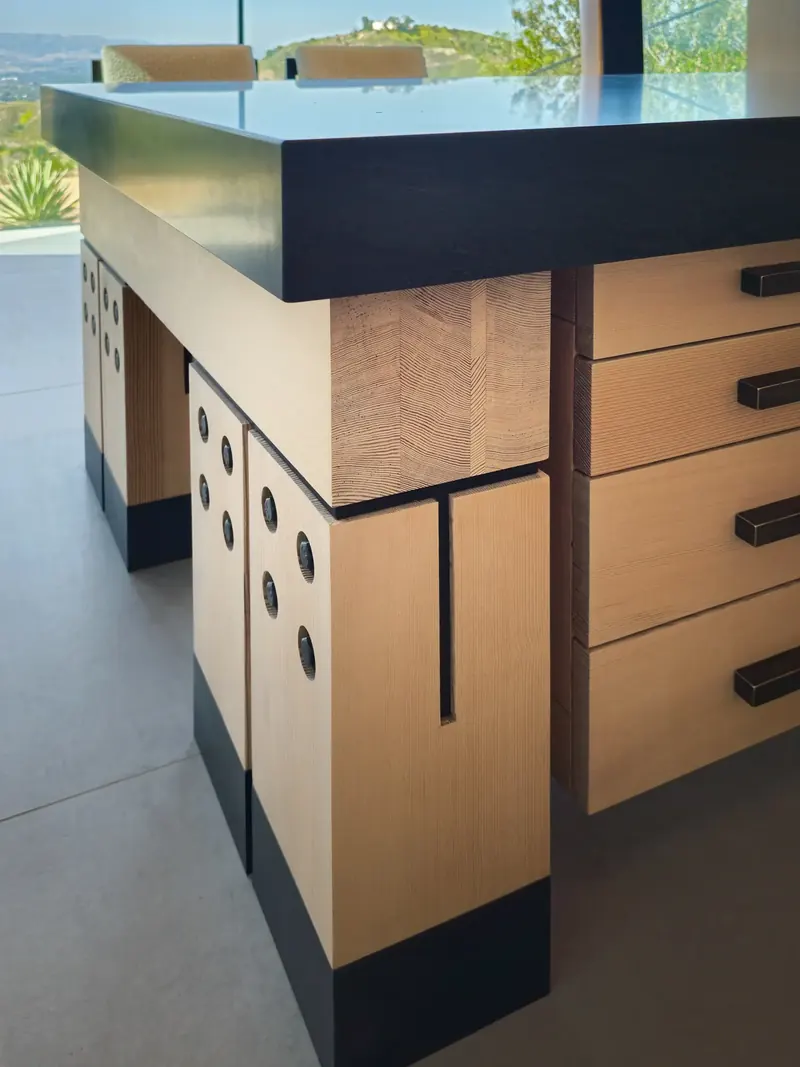 Corner of a heavy timber desk with a dark stone top, bolted timber legs with black bases and a bank of drawers
