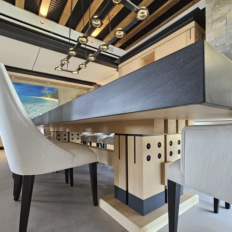 Long stone-topped dining table on heavy timber pedestals with bolted joints, beneath a beamed ceiling and linear pendant