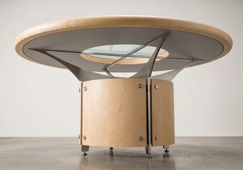 Low view under a round maple dining table, showing curved stainless steel supports above a cylindrical maple base