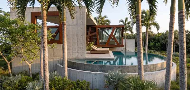 Circular concrete pool with a chaise on a concrete platform, seen through palm trunks, concrete wall and steel-framed volume beyond