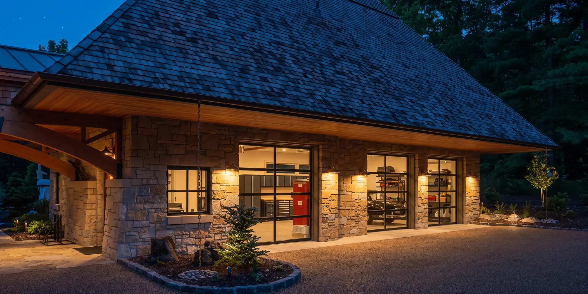 Stone garage at dusk with glass overhead doors, wall sconces and a deep shingle roof over a gravel court