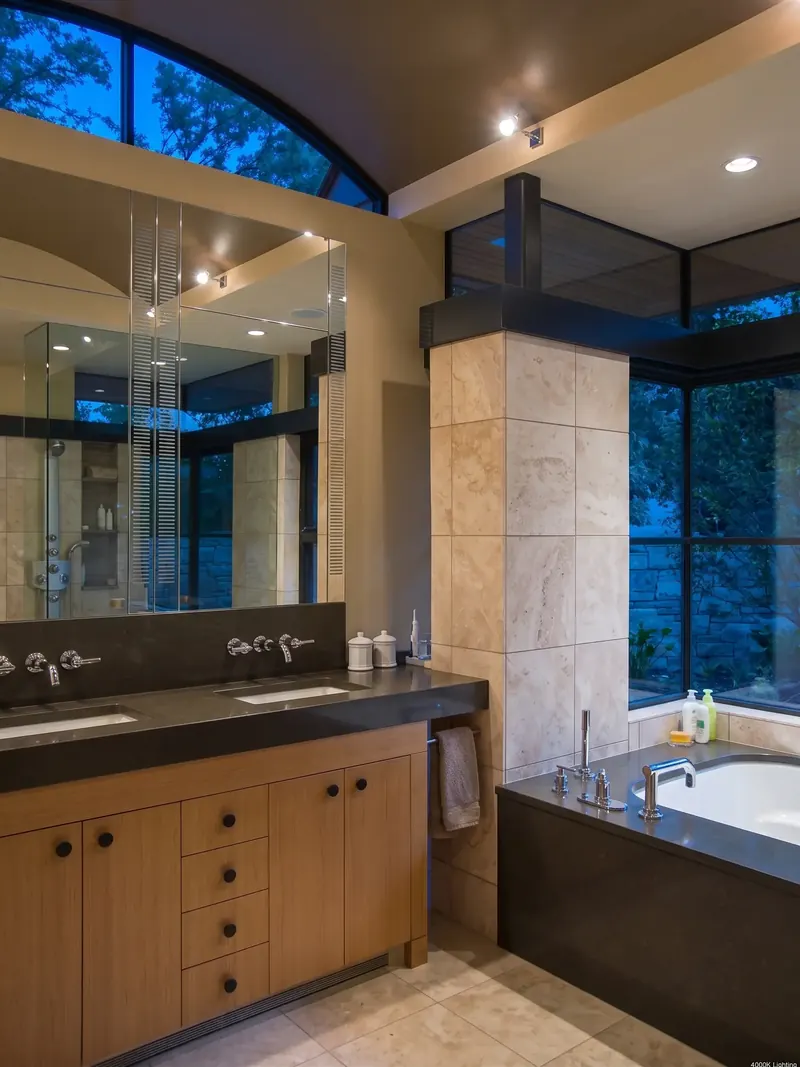 Bathroom at dusk with an arched window, wood double vanity, tiled column and a tub beside corner glass