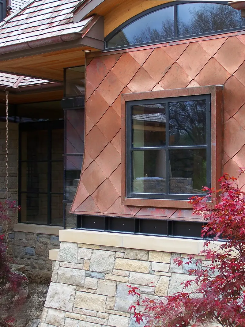 Flared wall of diamond-pattern copper panels around a dark-framed window, set on a stone base with red maple leaves