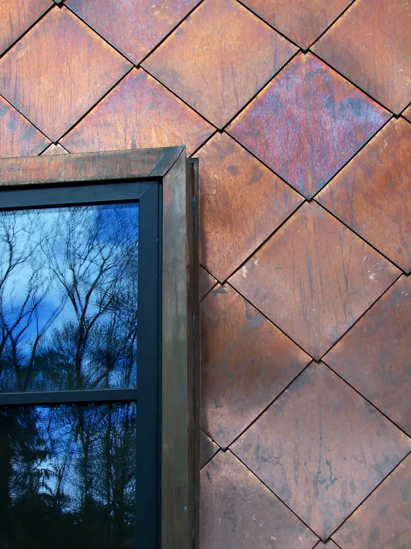 Close view of weathered copper diamond panels beside a window reflecting bare branches and blue sky