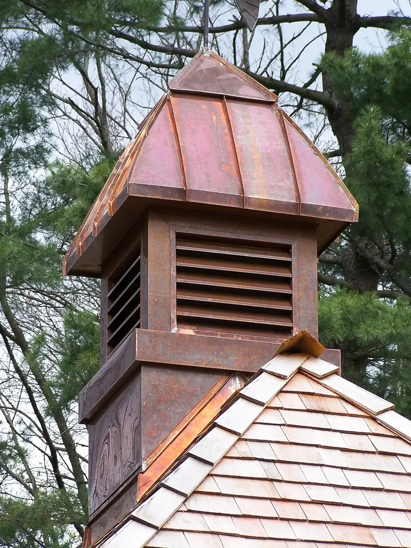 Louvered copper cupola with a standing-seam cap rising from a wood shingle roof against pine trees