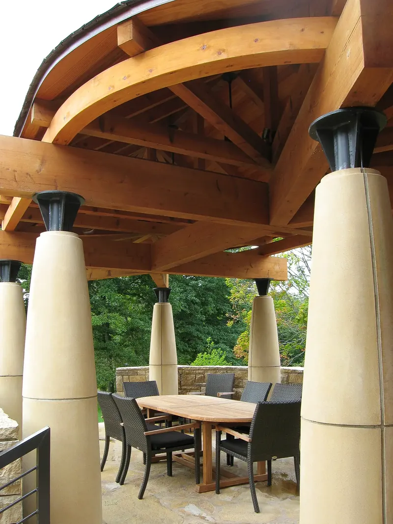 Curved and crossing timber beams resting on dark metal caps atop tapered columns above an outdoor dining table