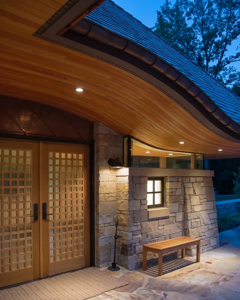 Entry at night, the lattice doors and stone wall lit beneath the curving wood soffit and glowing clerestory