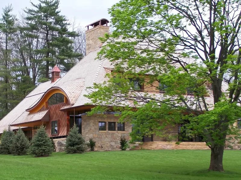 Sweeping shingle roof with a curved eyebrow dormer and cupola over a copper-clad bay and stone base, framed by trees