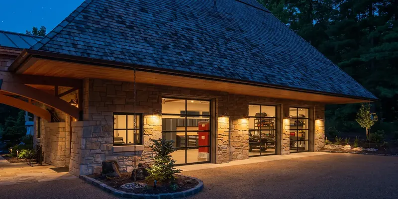 Stone garage at dusk with glass overhead doors, wall sconces and a deep shingle roof over a gravel court