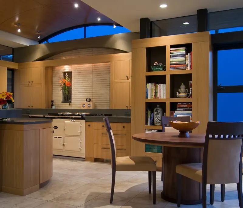 Kitchen at dusk with a cream cast iron stove under an arched clerestory, open bookshelves and a round table