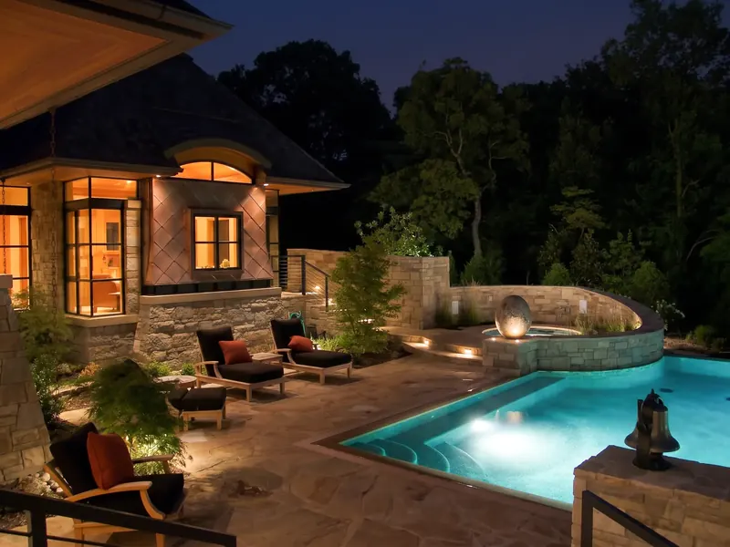 Pool terrace at night with lit lounge chairs, a curved stone spa wall and an illuminated egg sculpture