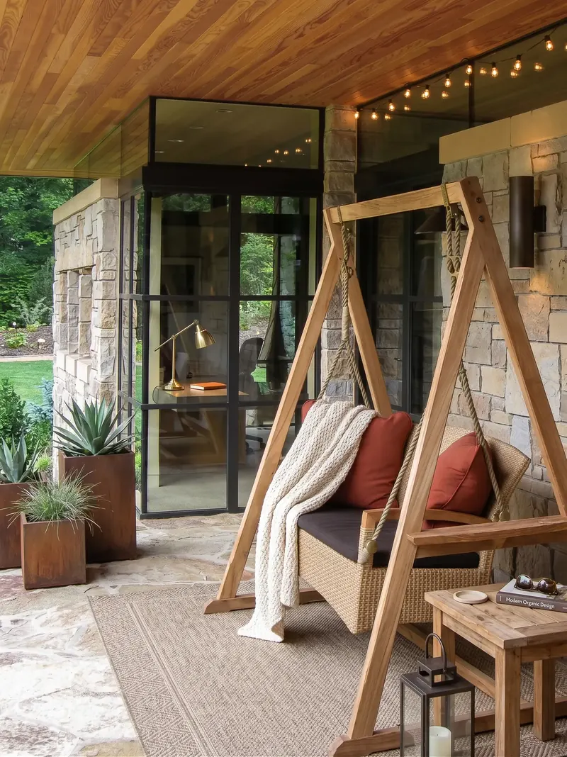 Covered porch with a wicker swing in a wood frame, stone walls, string lights and a glass-walled corner room with a desk