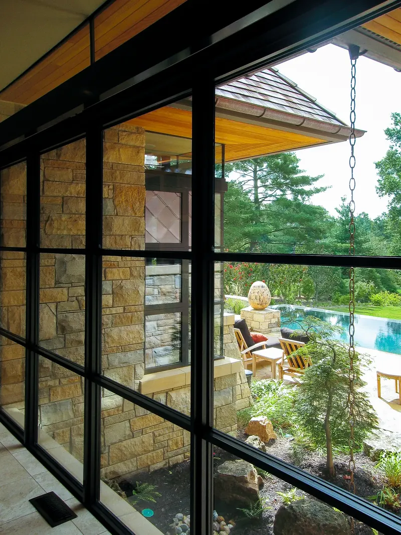 View through a dark-framed window wall to stone walls, a small cutleaf tree and the pool terrace beyond