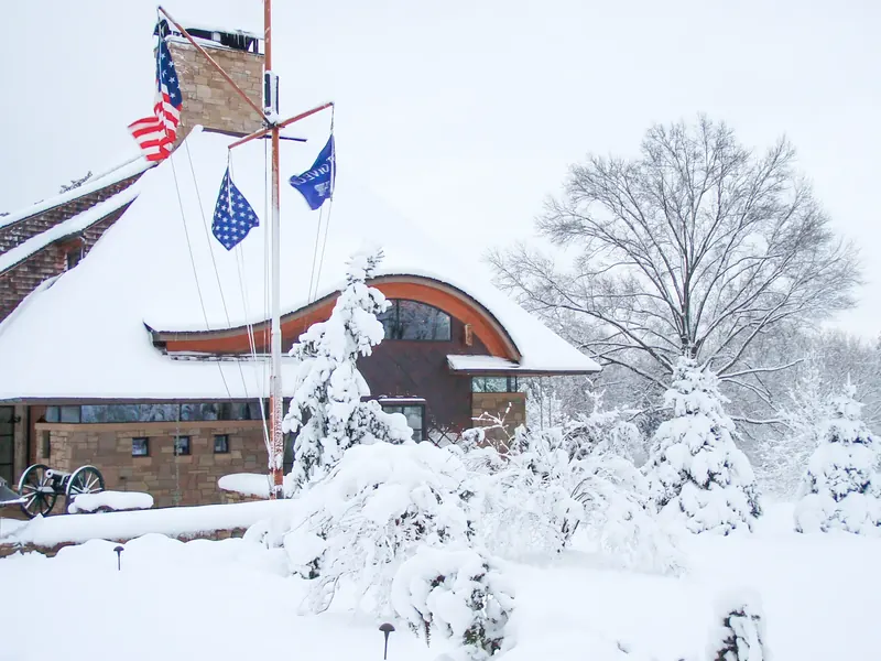 The curved roofline and copper-clad wall under heavy snow, with flags on a mast and snow-laden evergreens