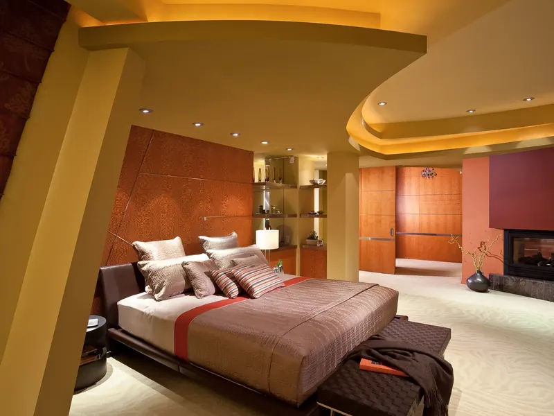 Bedroom under a curving lit ceiling, wood-paneled headboard wall, angled column and a fireplace at right