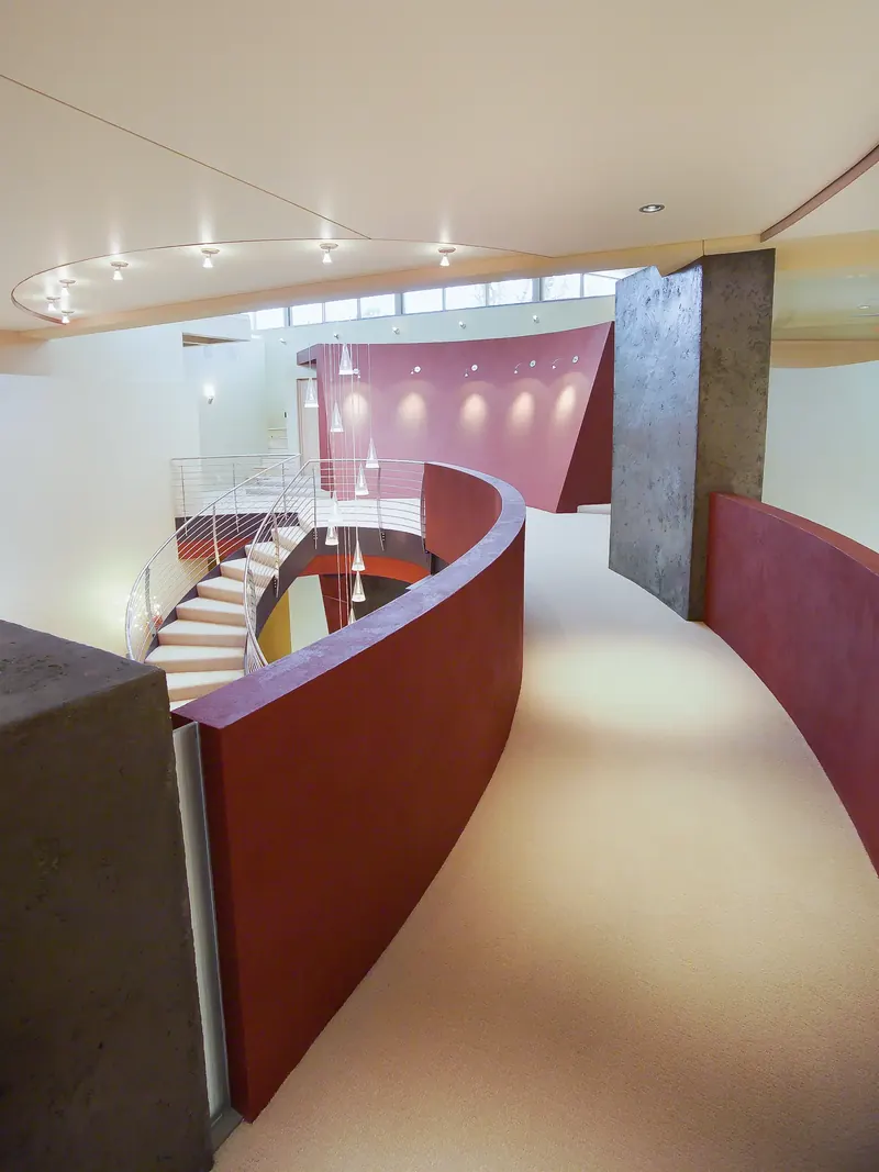 Carpeted curved bridge lined by red plaster walls, overlooking the staircase and pendant lights below