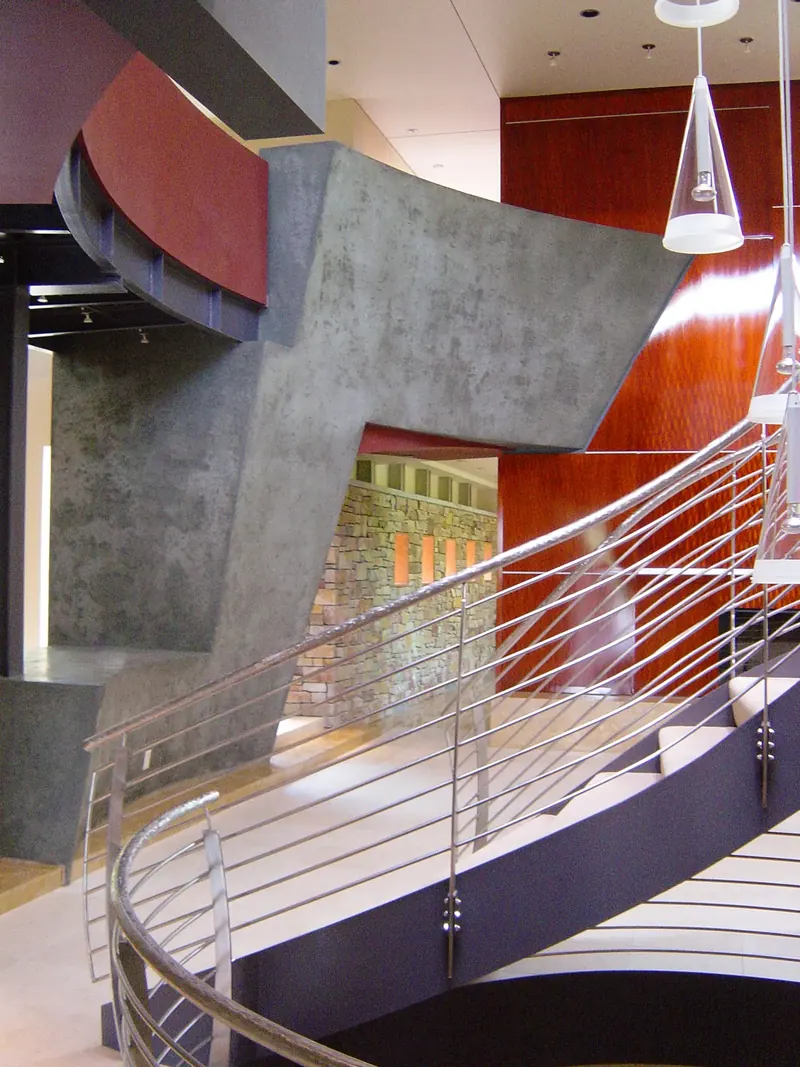 Angled grey plaster wall framing a view of a stone wall with lit niches, steel stair railing in the foreground