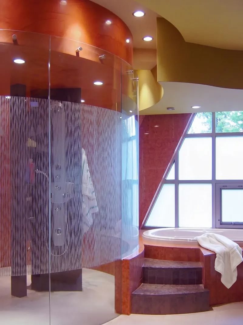 Round shower enclosed in curved etched glass beside a stepped tub and an angled window wall