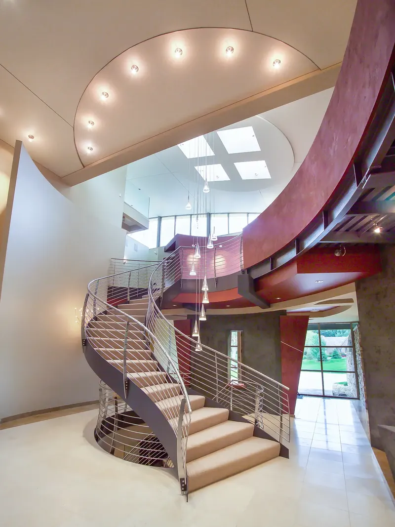 Sweeping carpeted staircase with steel rod railings rising beneath skylights and cone pendants toward a curved red bridge