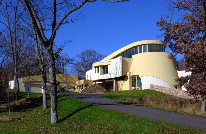 Daytime view up the driveway to the yellow sculptural house, garage wing at left and stone retaining walls