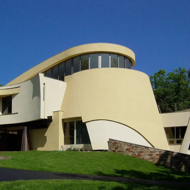 Tapered yellow stucco drum with a sweeping curved roof band of windows and a stone wall, under a deep blue sky