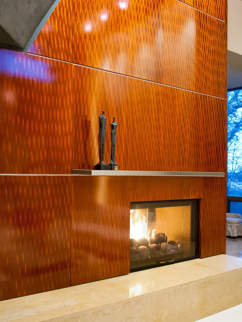 Close view of the fireplace: textured orange panels, steel mantel with two slender sculptures, stone hearth