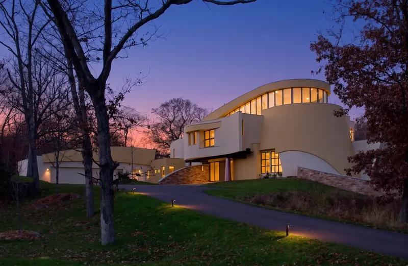 Curves and Colors House at dusk, its tapered yellow drum and curved band of windows glowing above a lit driveway