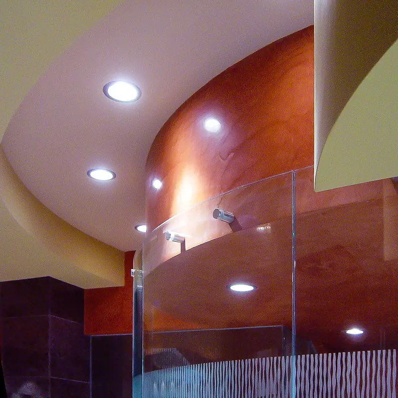 Top edge of a curved glass shower with steel fittings below a sweeping ceiling and a rust-colored curved wall