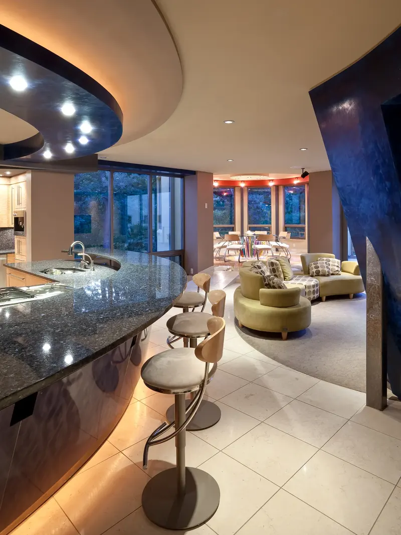 Curved stone bar and stools leading to a family area with green sofas and a round, windowed dinette