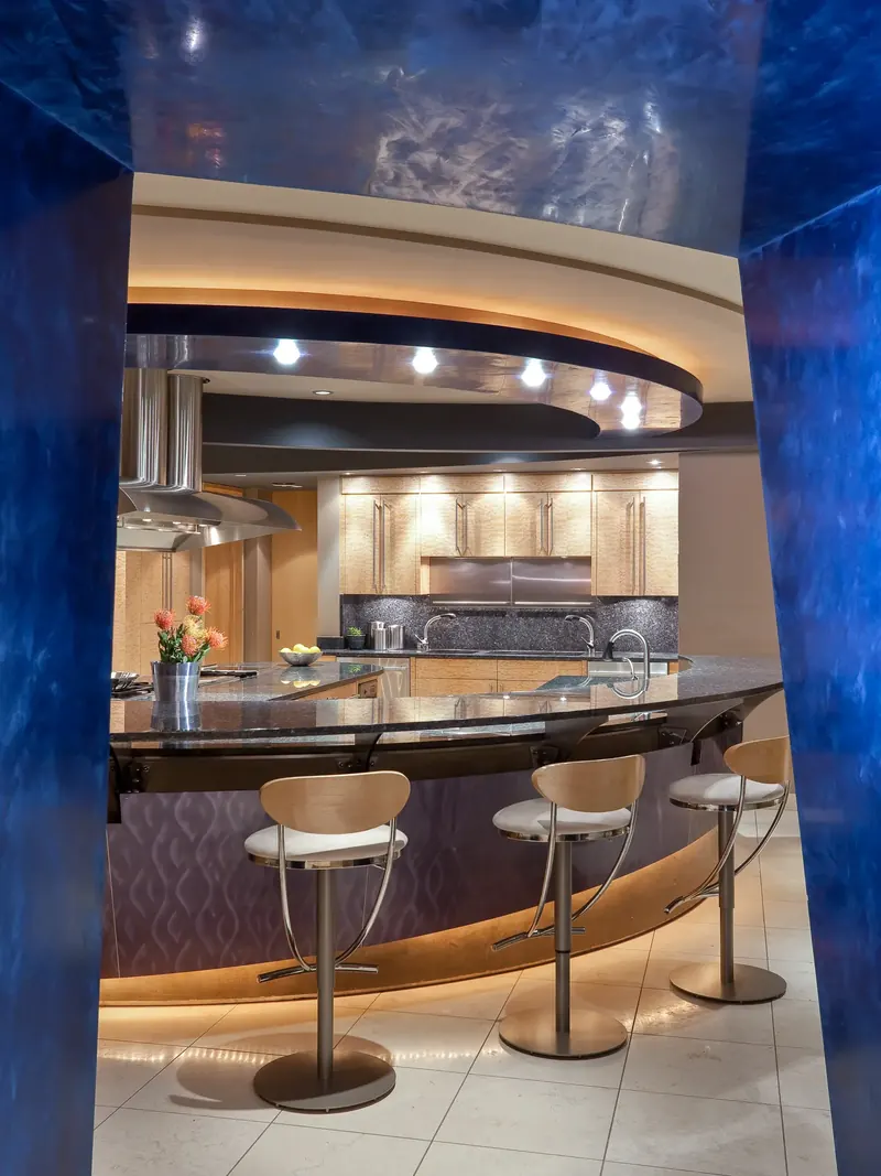 Curved island and three stools framed by blue Venetian plaster walls, light wood cabinets beyond